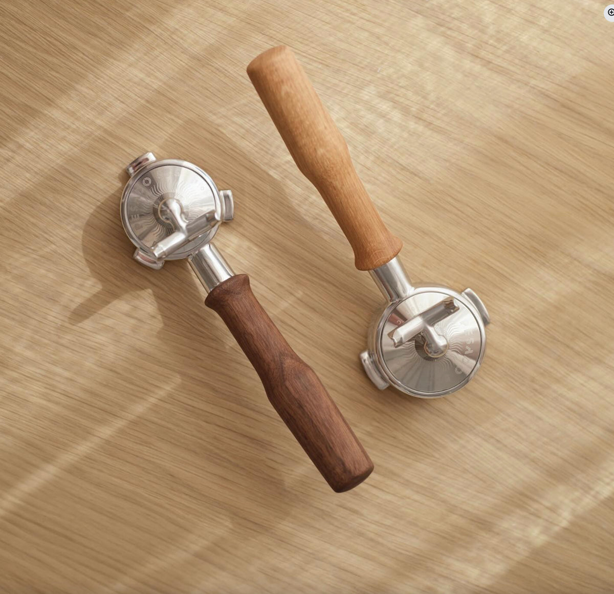 Två portafilter med handtag i valnöt och ek ligger på en ljus träyta i naturligt ljus.