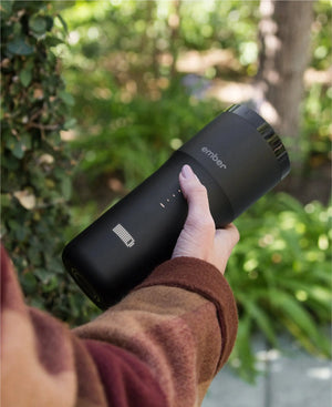 Electric Coffee Travel Mug black V2 - 355ml held in hand outdoors with lush green background.