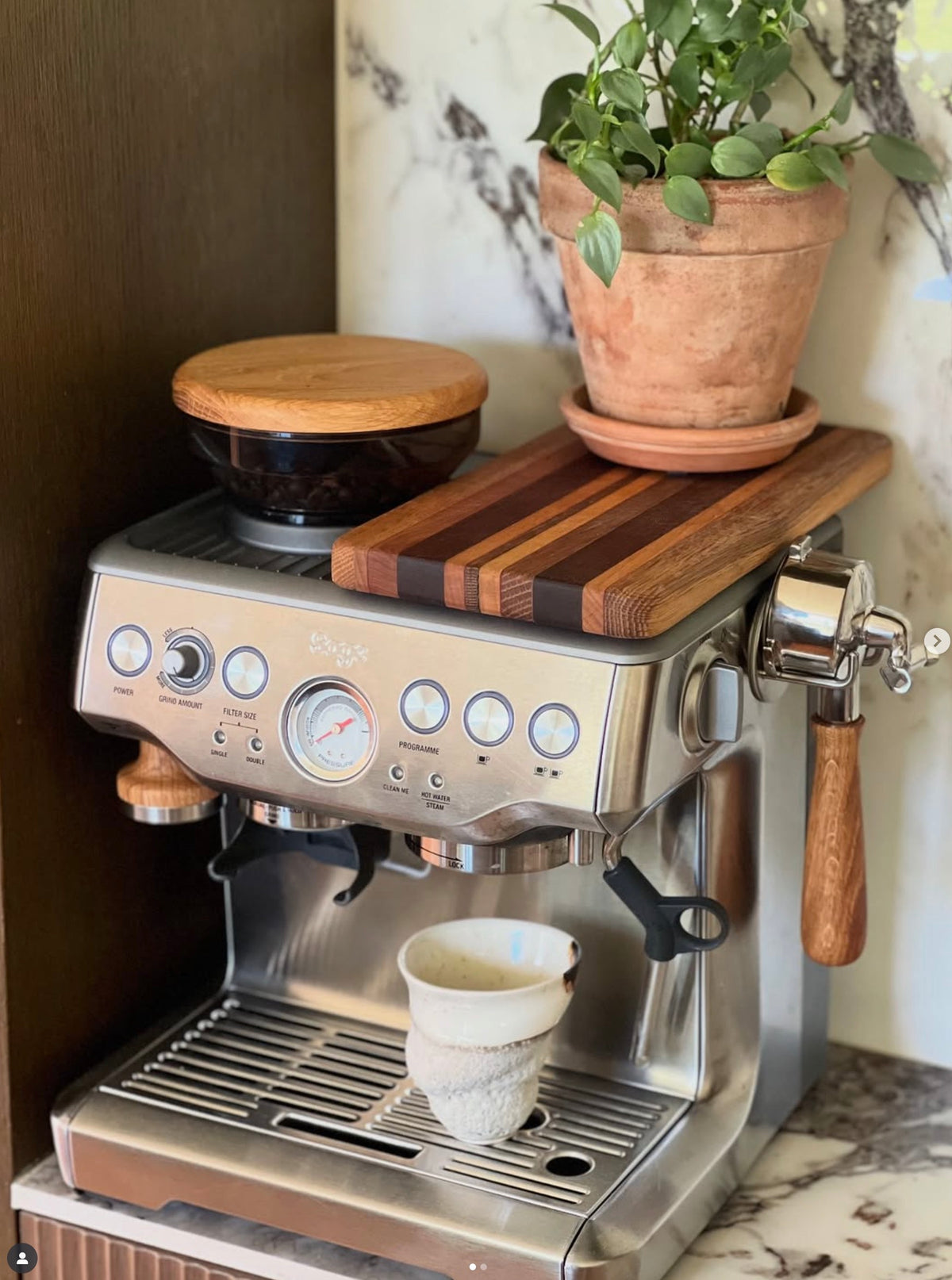 Espressomaskin i rostfritt stål med trädetaljer, kaffekvarn och växt på träskärbräda ovanpå.